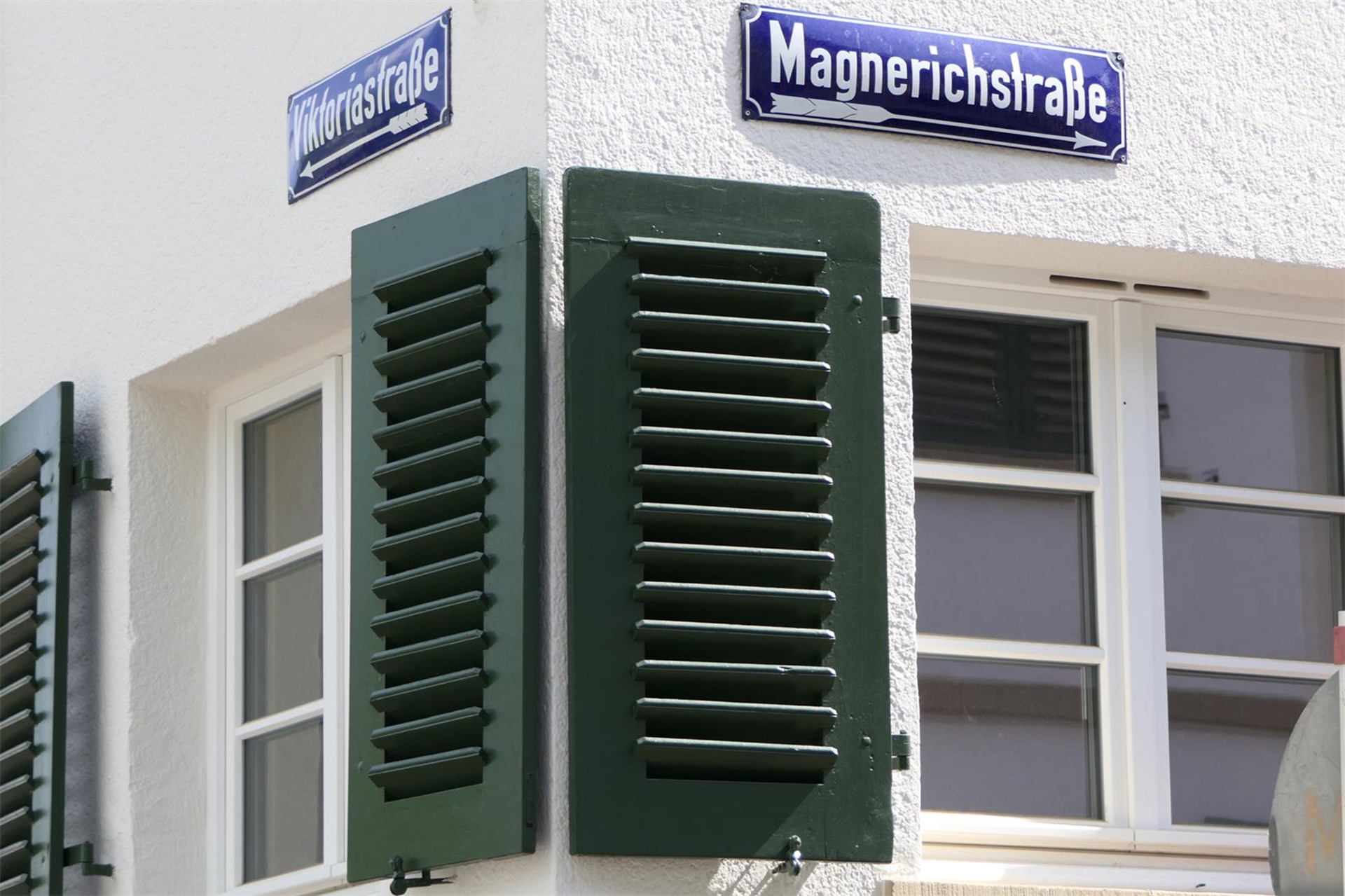 Blick auf Gebäudeecke; jeweils ein Fenster mit Fensterläden an beiden Hausseiten zu erkennen; zudem die an den Mauern angebrachten Straßenschilder „Viktoriastraße“ und „Magnerichstraße".