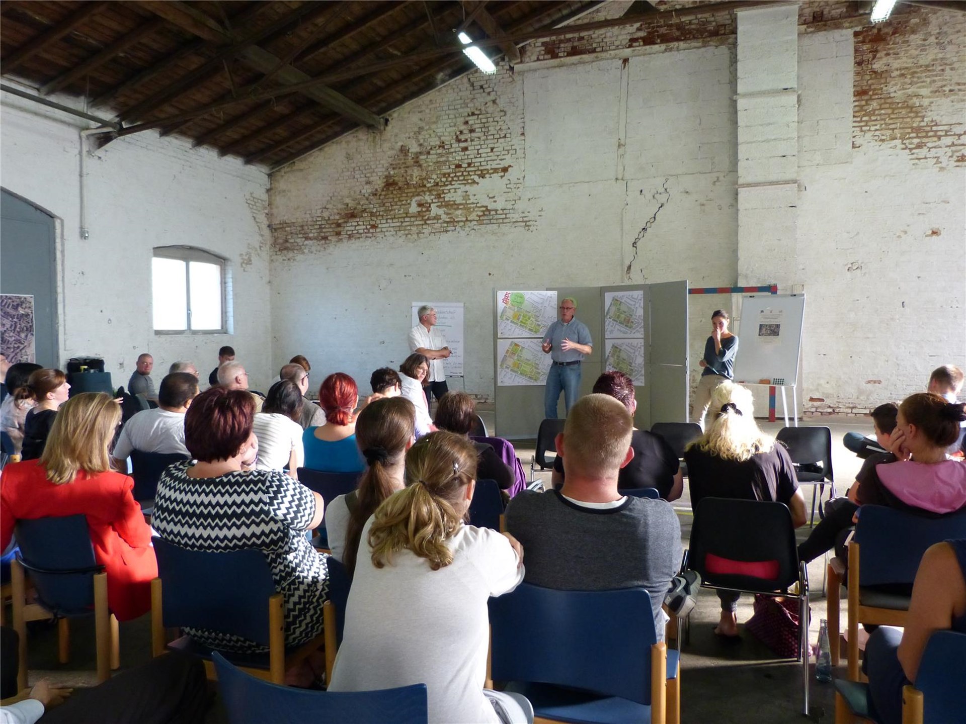 Viele Menschen sitzen auf Stuhlreihen und schauen nach vorne, wo drei weitere Menschen vor Schautafeln mit Plänen stehen.