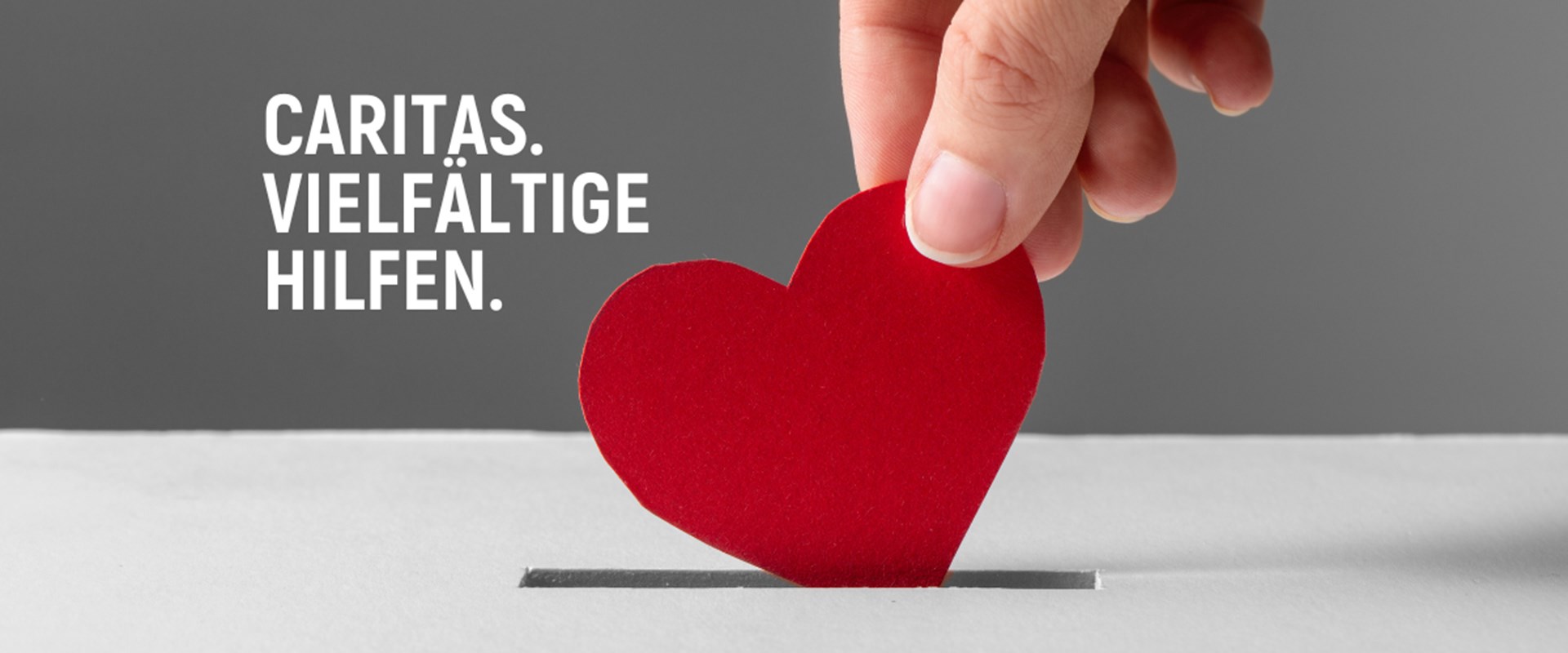 Symbolbild: Eine Hand, die ein rotes Papierherz in einen Geldschlitz steckt. Schriftzug: Caritas. Vielfältige Hilfen.